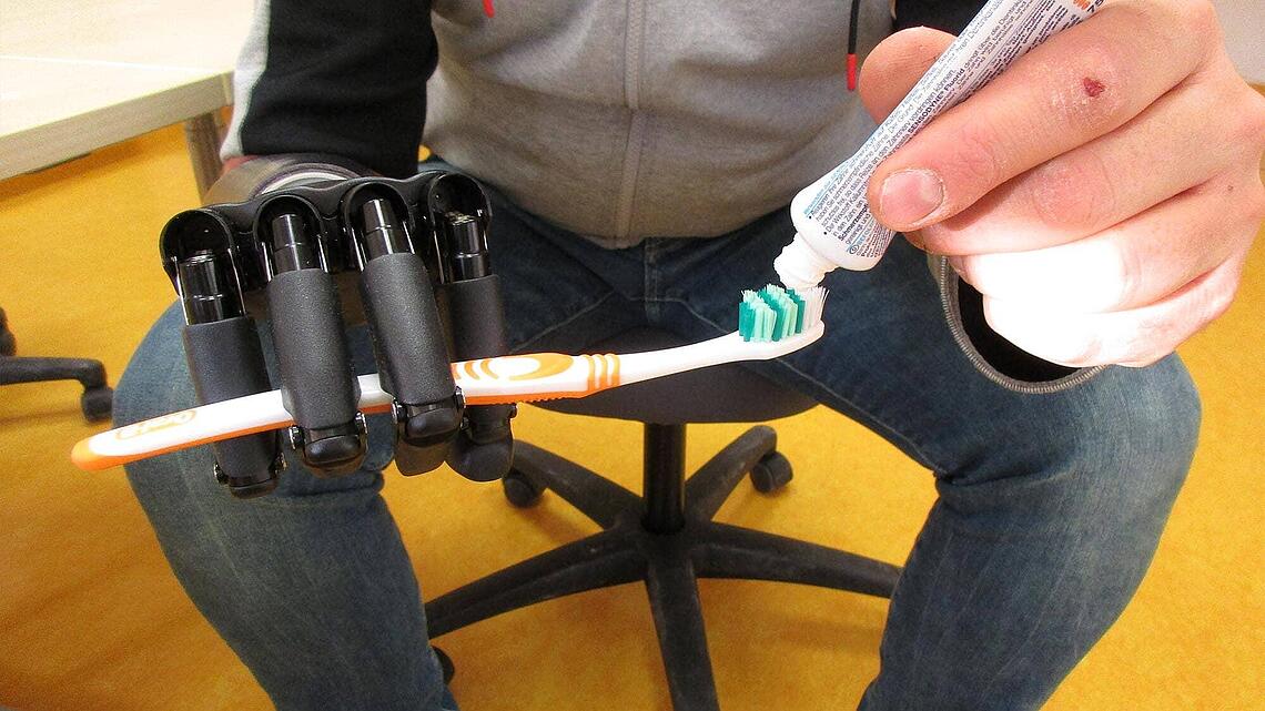 Mann sitzt auf Stuhl und hat an rechter Hand Prothese, mit linker Hand gibt er Zahnpasta auf die Zahnbürste bei der Prothesen-Gebrauchsschulung