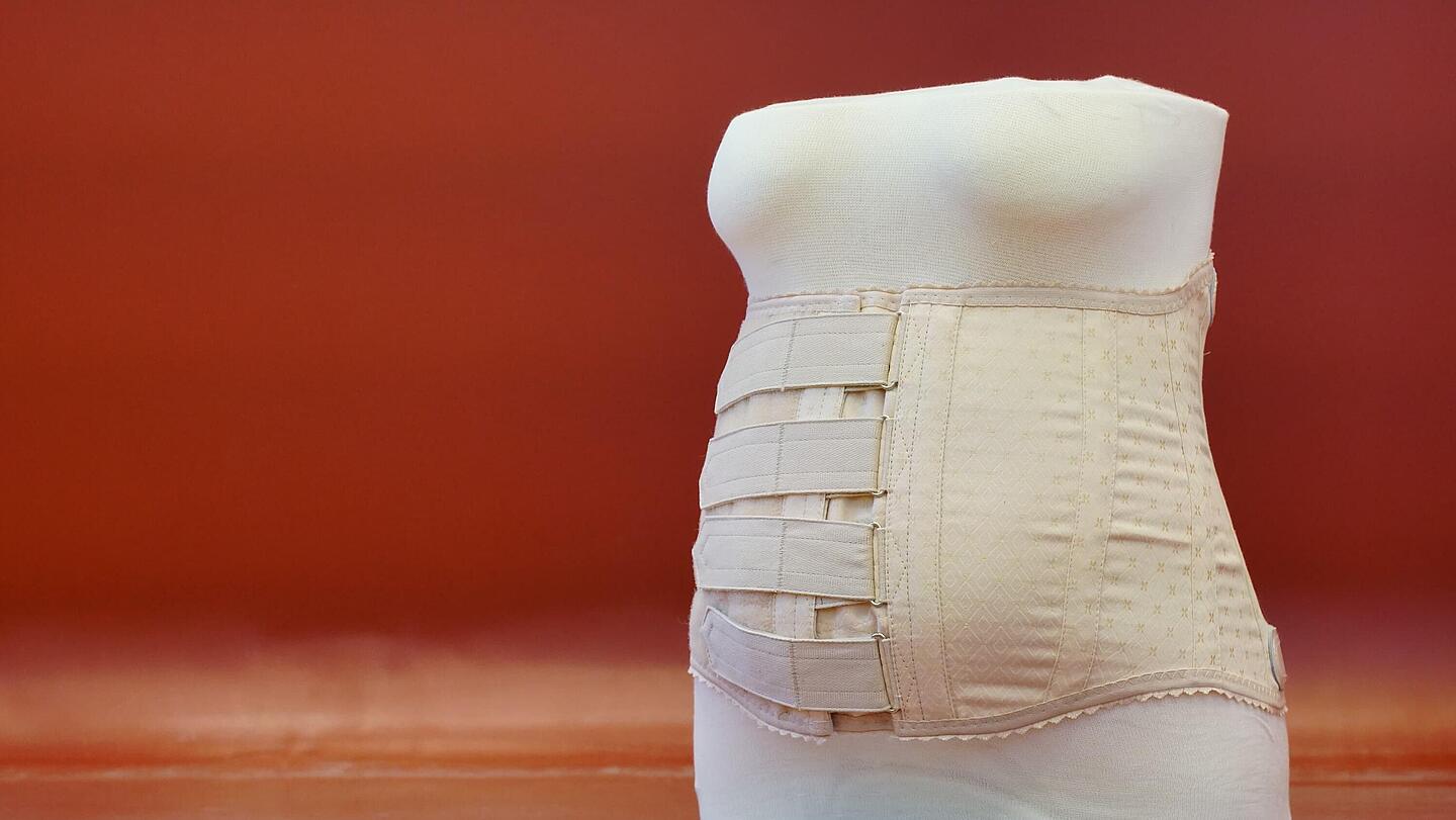 Vorderansicht eines Stützmieders nach Schanz mit vertikal verlaufenden Metallverstärkungsstäben vor rotem Hintergrund